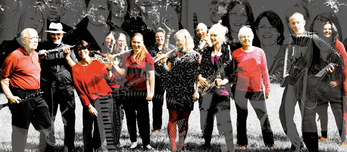 Die 13 Musiker:innen des Orchesters mit Instrumenten, in rot und schwarz gekleidet